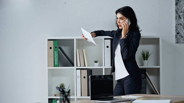 Bizneswoman rozmawia na smartfonie i trzyma papier w pobliżu laptopa na zamazanym pierwszym planie w biurze  - Zdjęcie, obraz