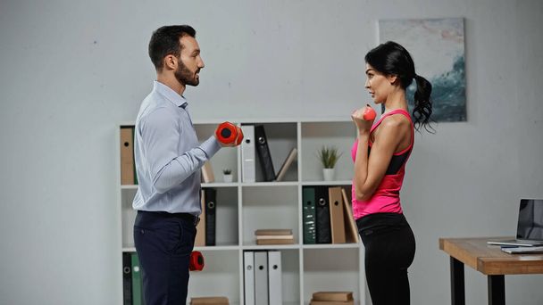 Side view of businessman training with fitness trainer and dumbbells in office  - Photo, image