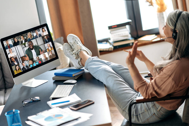 Verkkotiedotustilaisuus. Moderni ikäinen harmaa tukka nainen kommunikoi työtovereiden kanssa verkossa videopuhelun kautta käyttää sovellusta ja tietokonetta, istuu hänen kotitoimistossaan, heittää jalkansa pöydälle. - Valokuva, kuva