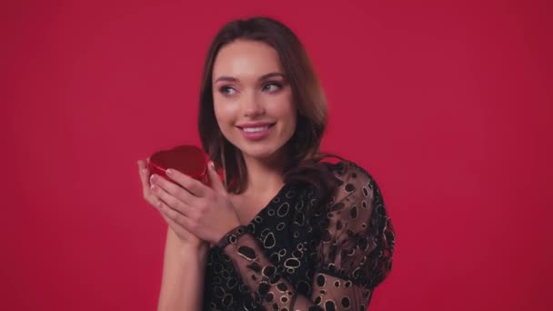 mujer feliz en vestido negro sosteniendo caja en forma de corazón aislado en rojo - Imágenes, Vídeo
