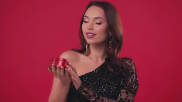 χαρούμενη γυναίκα με μαύρο φόρεμα ανοίγοντας κουτί δώρου σε σχήμα καρδιάς απομονωμένο σε κόκκινο - Πλάνα, βίντεο
