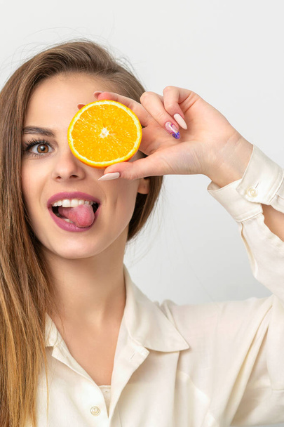 Retrato de una alegre mujer joven caucásica que cubre el ojo con una rebanada de naranja y saca la lengua lleva camisa blanca sobre un fondo blanco - Foto, Imagen