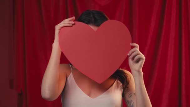 χαρούμενη γυναίκα που καλύπτει το πρόσωπο με χάρτινη καρδιά και χαμογελά στο κόκκινο - Πλάνα, βίντεο