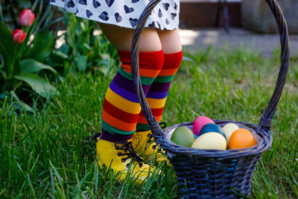 Nahaufnahme der Beine von Kleinkind Mädchen mit bunten Strümpfen und Schuhen und Korb mit bunten Eiern. Kinder vergnügen sich bei der traditionellen Ostereiersuche an warmen, sonnigen Tagen im Freien. - Foto, Bild