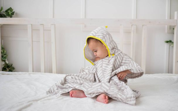 Baby Junge sitzt seitlich in einem Kapuzenhandtuch auf einem weißen Bett - Foto, Bild