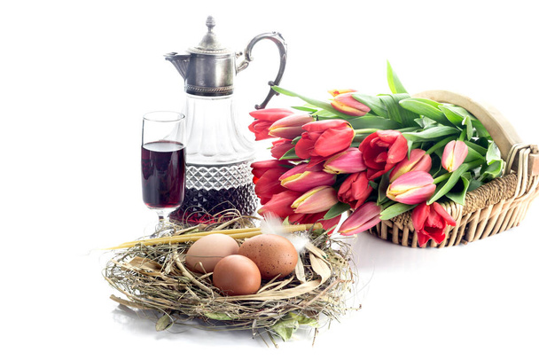 La composición de Pascua. Huevos, tulipanes rojos, velas de la iglesia y vino en una copa y una jarra sobre fondo blanco de cerca. - Foto, Imagen