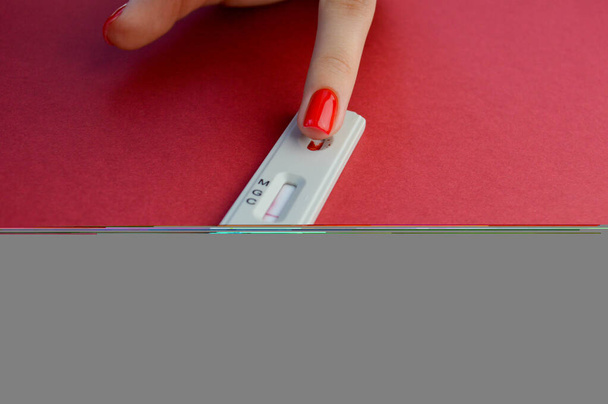 test op coronavirus ligt op een rode achtergrond. Een meisje met een rode manicure brengt een doorboorde vinger aan om bloed te nemen. gevoelige test op coronavirusantilichamen. - Foto, afbeelding
