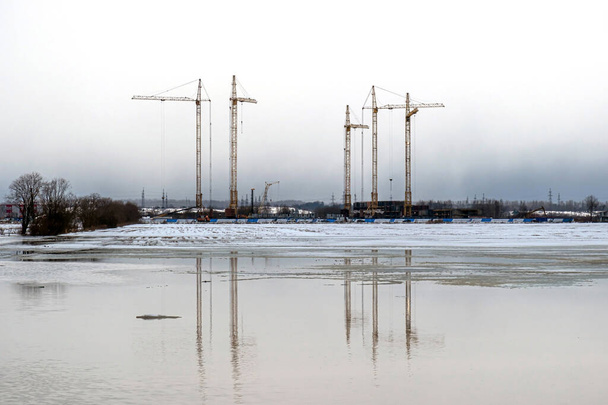 Dźwigi budowlane na skraju pola zalane topniejącą wodą. Przedmieścia Sankt Petersburga. Koniec zimy, gołe drzewa. - Zdjęcie, obraz