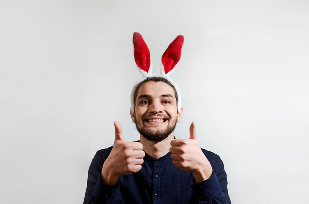 Okouzlující mladý muž s vousy v košili v kostýmu velikonočního zajíčka. Muž s králičíma ušima. Osoba se zaječím ušima se usměje a zvedne dva prsty. Tvůrčí prapor na oslavu Velikonoc. - Fotografie, Obrázek