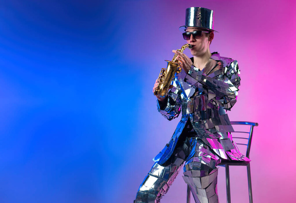 a male artist in a mirrored suit and a hat with a saxophone in neon light - Valokuva, kuva