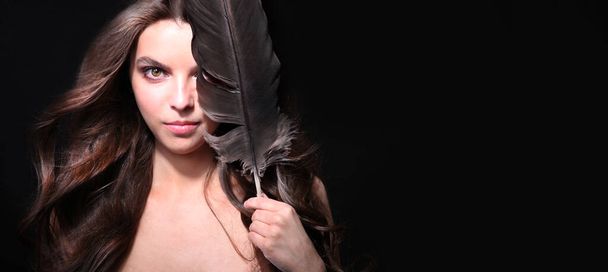 Una hermosa mujer mística con una pluma de cuervo en la mano y pelo que fluye sobre un fondo negro - Foto, imagen