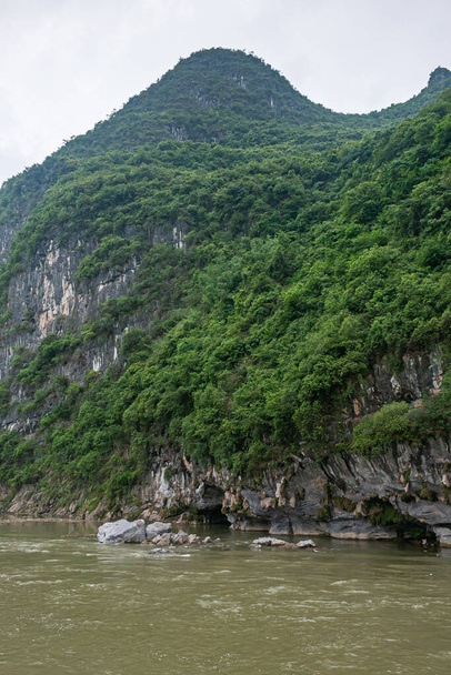 Guilin, Çin - 10 Mayıs 2010: Li Nehri boyunca. Koyu kayalıklardaki yeşil su seviyesindeki mağaralar kısmen yeşil ağaçlarla kaplı gümüş gökyüzünün altındaki yüksek dağlar.. - Fotoğraf, Görsel