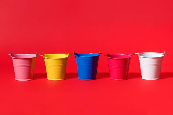 Cinco baldes de brinquedo multicoloridos em um fundo vermelho. Amarelo azul branco rosa e balde vermelho. Conceito de renovação futura ou início da colheita ou plantação de culturas - Foto, Imagem