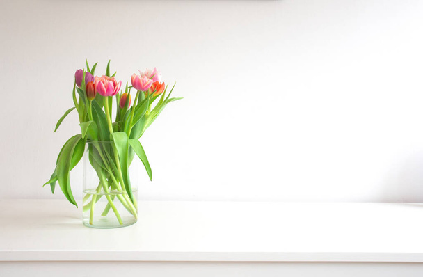 Čerstvé světlé kytice barevných tulipánů u bílé stěny na dřevěné polici ve skleněné váze s prostorem pro text, jaro, duben, dekorace interion koncept moderní design. Kopírovat prostor - Fotografie, Obrázek