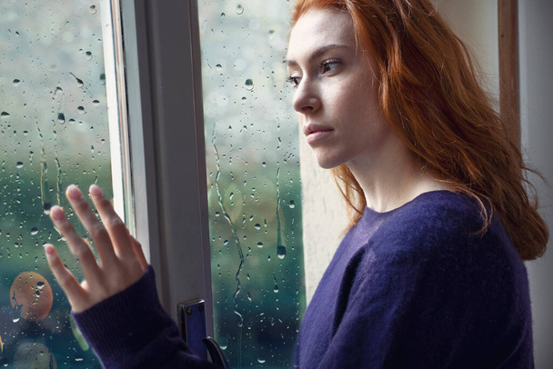 Portret van een bezorgd meisje dat thuis door het raam kijkt - Foto, afbeelding