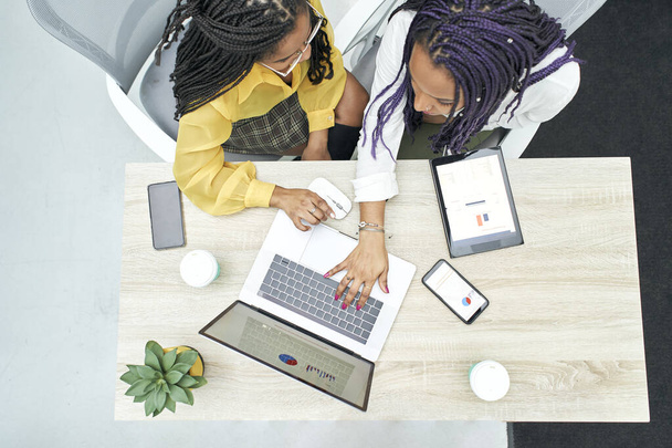 Vista superior de dos jóvenes mujeres negras que trabajan en la oficina. Revisión de datos analíticos en varios dispositivos electrónicos. Vista de Zenith. - Foto, imagen