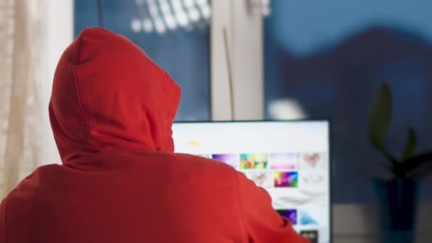 Muž v kapuci pracuje večer doma za monitorem počítače, pohled zezadu - Záběry, video