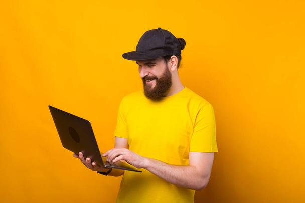 Jovem barbudo feliz usando laptop sobre fundo amarelo - Foto, Imagem