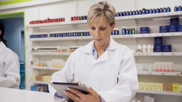 Caucasian female pharmacist scrolling through scripts on digital tablet smiling wearing coat in clean pharmacy  - Footage, Video