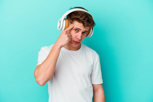 Joven hombre caucásico escuchando música con auriculares aislados sobre fondo azul apuntando templo con el dedo, pensando, centrado en una tarea. - Foto, imagen