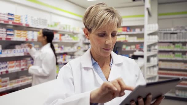 farmacista caucasica femminile digitando su tablet digitale e-mail script ai clienti che indossano cappotto bianco in piedi in farmacia  - Filmati, video