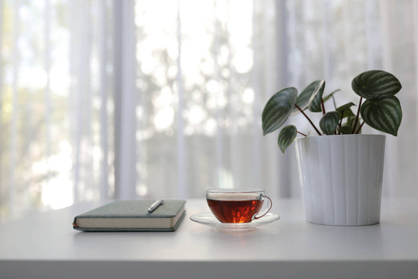 punainen tee läpinäkyvässä lasissa kasvi ja muistikirja ja valkoinen pöytä nähdä läpi verho - Valokuva, kuva