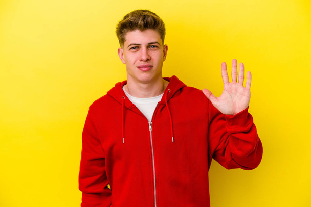 Nuori valkoihoinen mies eristetty keltaisella taustalla hymyilevä iloinen osoittaa numero viisi sormin. - Valokuva, kuva