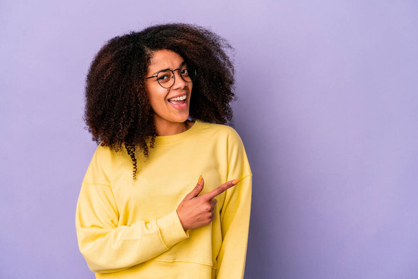 Jeune femme frisée afro-américaine isolée sur fond violet souriant et pointant du doigt, montrant quelque chose à vide. - Photo, image