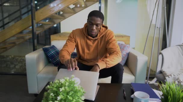 Νεαρός αφροαμερικανός επιχειρηματίας φοιτητής ελεύθερος επαγγελματίας χρησιμοποιώντας συσκευή laptop ακουμπώντας στον καναπέ στο γραφείο στο σπίτι - Πλάνα, βίντεο