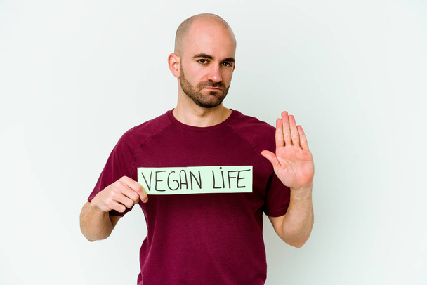 Joven hombre calvo caucásico sosteniendo un cartel de vida vegana aislado sobre fondo púrpura de pie con la mano extendida mostrando señal de stop, lo que le impide. - Foto, Imagen