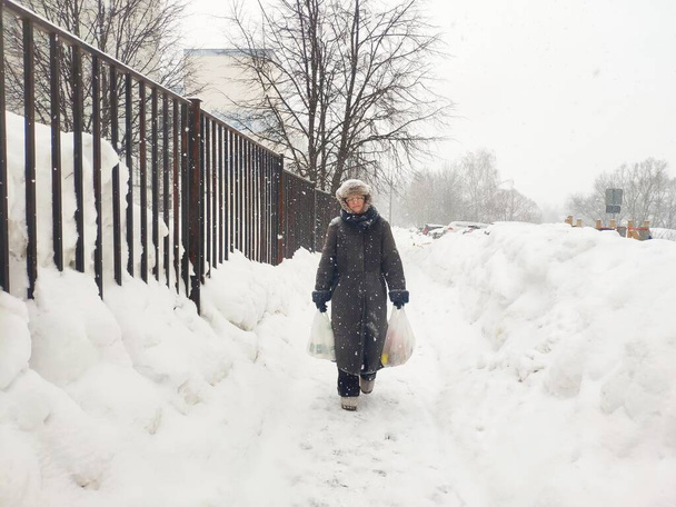 Yaşlı olgun bir kadın yoğun bir kar yağışında alışveriş torbaları taşır. Orta yaşlı bir büyükanne kar fırtınasında yürüyor. Şehirdeki kötü hava kavramı - Fotoğraf, Görsel