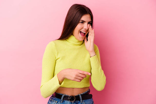Young caucasian woman isolated on pink background saying a gossip, pointing to side reporting something. - Foto, immagini