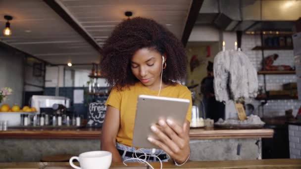 Kulaklık takan güzel bir yarış kadını kafede oturmuş dijital tablet kullanarak arkadaşlarıyla görüntülü konuşma yapıyor.  - Video, Çekim