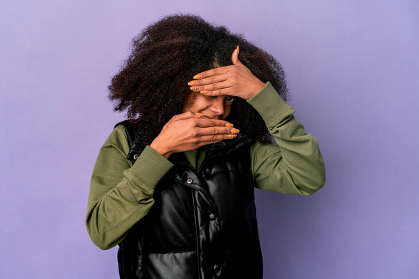 Giovane donna riccia africana americana isolata su sfondo viola lampeggia alla fotocamera attraverso le dita, imbarazzato volto di copertura. - Foto, immagini