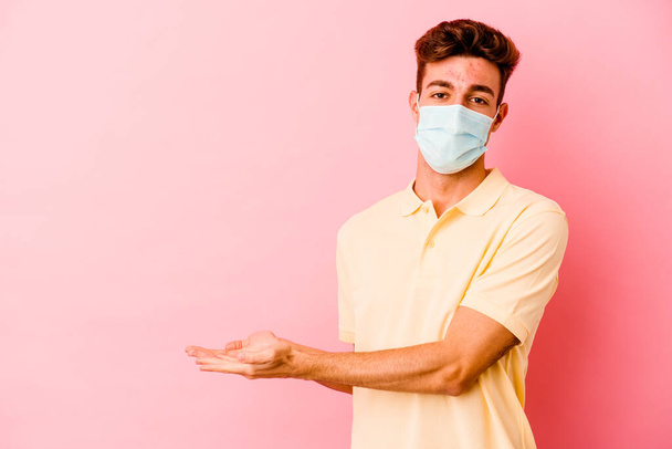 Junger kaukasischer Mann trägt einen Coronavirus-Schutz isoliert auf rosa Hintergrund und hält einen Kopierraum auf einer Handfläche. - Foto, Bild