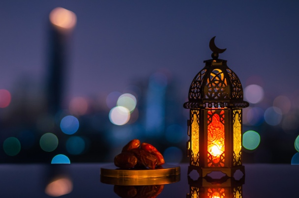 Lanterna que tem símbolo da lua em cima e pequeno prato de frutas datas com céu noturno e cidade bokeh fundo de luz para a festa muçulmana do mês santo do Ramadã Kareem. - Foto, Imagem