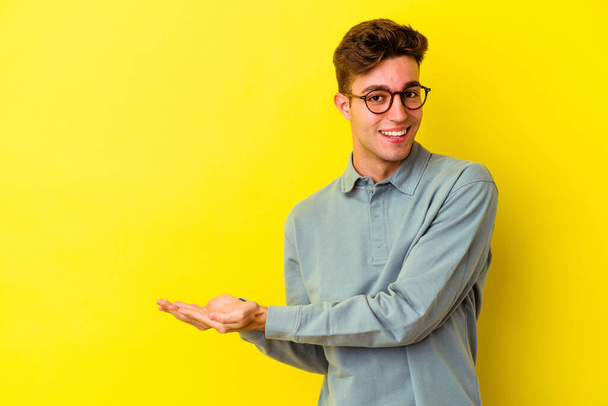 Jeune homme caucasien isolé sur fond jaune tenant un espace de copie sur une paume. - Photo, image