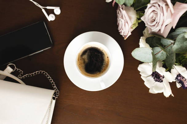 Una tazza di caffè con un bouquet di tenero anemone, rami di eucalipto e rose viola pastello, una borsa donna, cuffie e un telefono su un tavolo di legno marrone. Sfondo sfocato - Foto, immagini