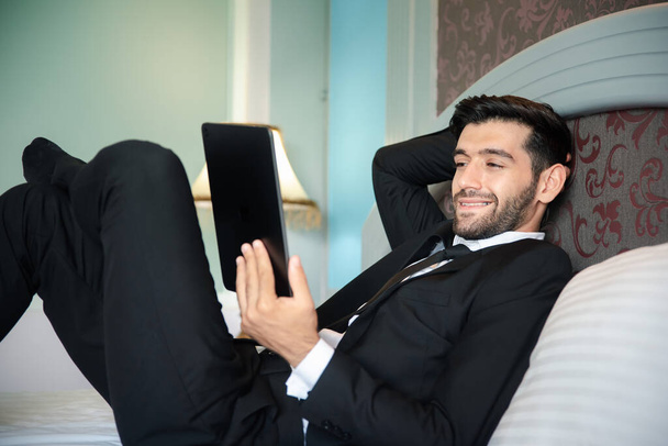 Handsome young bearded businessman lying on bed with tablet - Foto, imagen