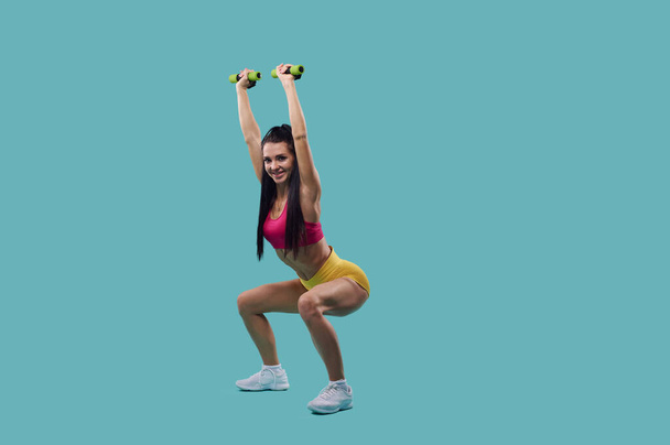 Woman with beautiful body performs dumbbell squats isolated on blue background with copy space. - Photo, Image