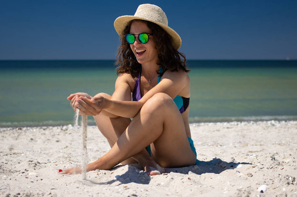 Spannende jonge vrouw die op het strand speelt, zand uit de handen loslaat, een zomerse strohoed, een zonnebril en een blauwe bikini draagt, geniet van de zomervakantie zittend met een blauwe oceaan achtergrond. - Foto, afbeelding