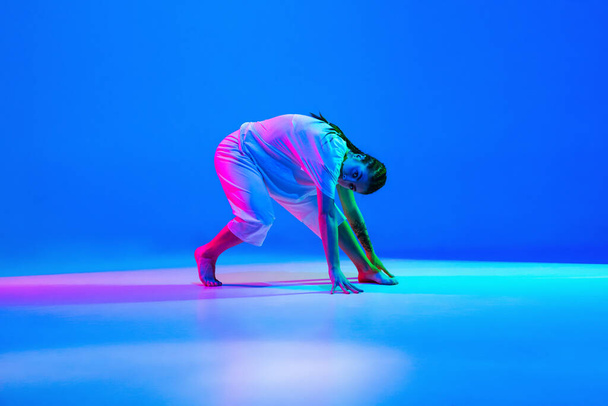 Sportliches Mädchen, das Hip-Hop in weißer Kleidung auf blauem Hintergrund in Neonlicht tanzt - Foto, Bild