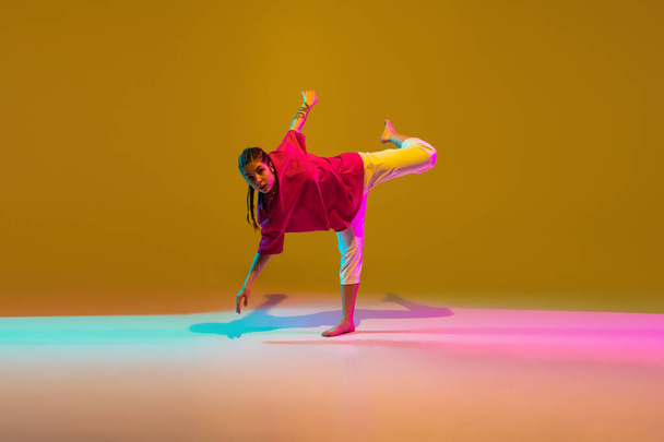 Gelukkig meisje danst hip-hop in stijlvolle kleding op kleurrijke achtergrond in neon licht - Foto, afbeelding