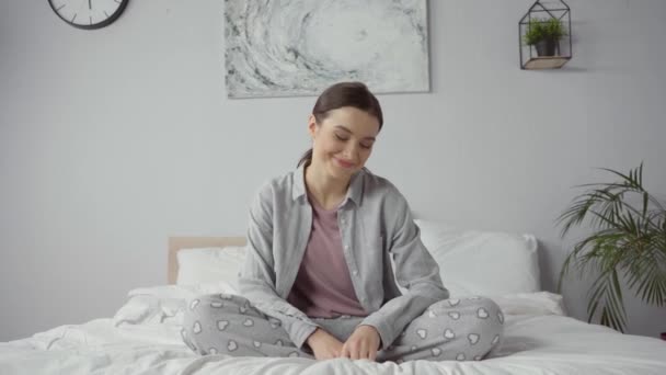 Χαρούμενη γυναίκα που κάθεται στο κρεβάτι και χαμογελάει στο σπίτι - Πλάνα, βίντεο