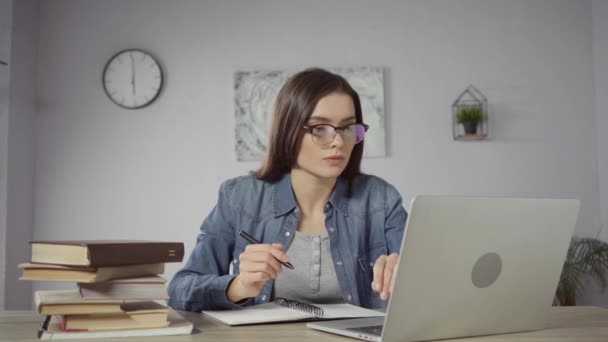 mujer joven con gafas escribiendo en un cuaderno mientras aprende en línea - Imágenes, Vídeo