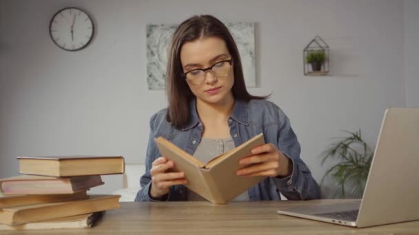 mujer joven en gafas libro de lectura cerca del ordenador portátil en el escritorio - Metraje, vídeo