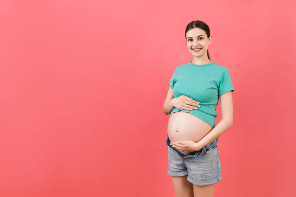 Die werdende Mutter in Jeans mit Reißverschluss zeigt ihren nackten Schwangerschaftsbauch vor buntem Hintergrund mit Kopierraum. Schwangerschaftskonzept. - Foto, Bild