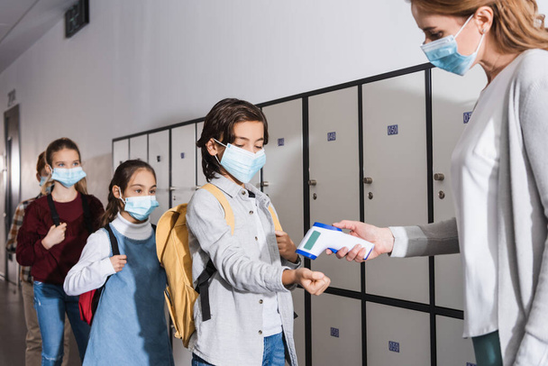 Iskolásfiú orvosi maszkban a tanár közelében áll, érintésmentes hőmérővel az iskolai folyosón.  - Fotó, kép