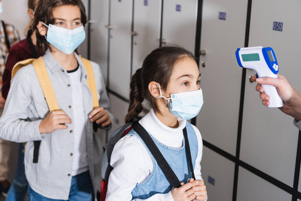 Μαθητής με ιατρική μάσκα στέκεται κοντά σε δάσκαλο μέτρησης της θερμοκρασίας στο διάδρομο του σχολείου  - Φωτογραφία, εικόνα