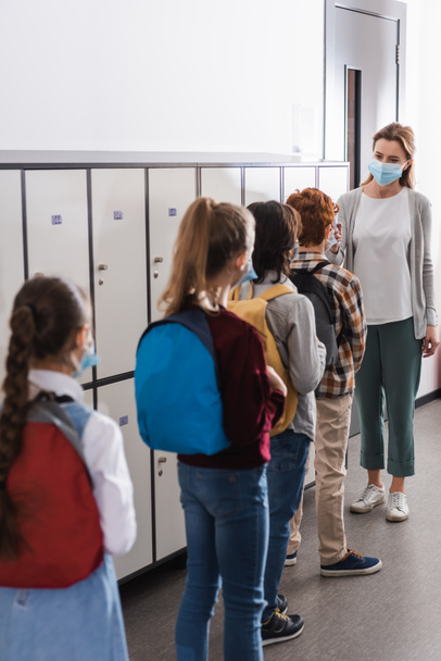 Δάσκαλος που κρατά θερμόμετρο χωρίς επαφή κοντά σε μαθητές με ιατρικές μάσκες σε θολή πρόσοψη στο διάδρομο  - Φωτογραφία, εικόνα